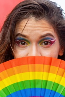 Close-up of a woman with vibrant rainbow makeup and jewels, holding a rainbow fan, symbolizing