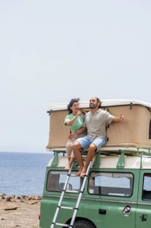 A blind man sitting atop a motorhome by the sea, accompanied by a female guide. They enjoy the