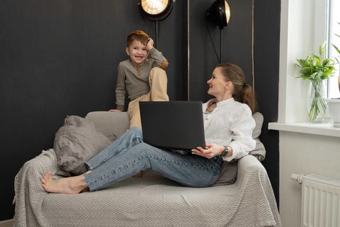 A mother lounges on a cozy sofa, working on her laptop, while her playful child sits nearby. The