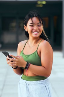 A cheerful teenager wearing a green top and gray sweatpants smiles while holding a phone. Strands