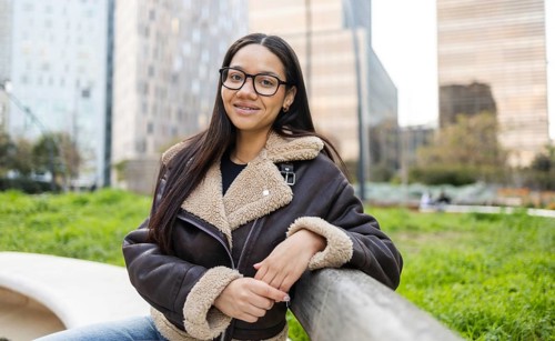 A hispanic woman in a cozy jacket stands confidently in an urban park, embodying a modern business
