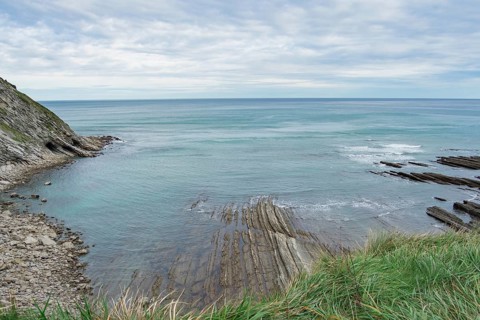 A serene coastal landscape showcasing a calm sea, striated rock formations, and lush green grass