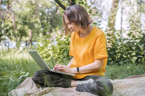 Woman sits cross-legged on a blanket in a lush park, using her laptop. Surrounded by greenery, she