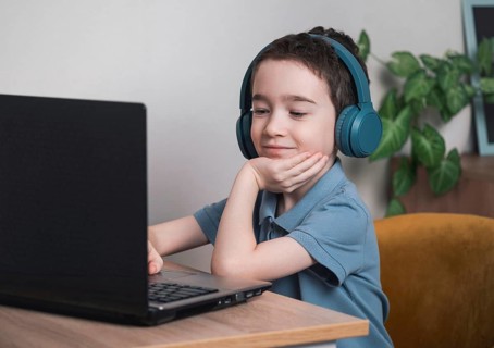 A young boy, smiling in headphones, engaged with a laptop. He is sitting at a wooden desk,