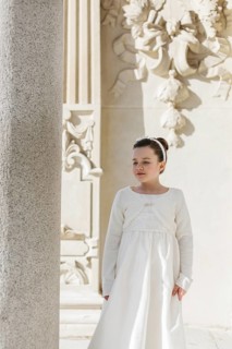 A young girl stands gracefully wearing a First Communion dress near a detailed stone pillar with