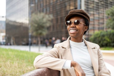 A fashionable black woman wearing sunglasses and a hat relaxes on a park bench in an urban area,