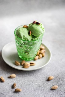 A scoop of creamy pistachio ice cream in a textured green glass sits on a plate, surrounded by
