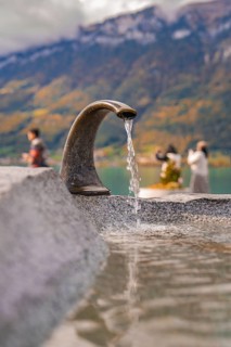 Fountain with running water, people at the lake with mountains in the background, Lake Brienz,