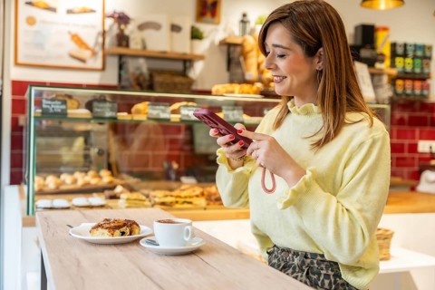 Smiling woman browsing her smartphone while relaxing at a bakery counter, enjoying a warm cup of
