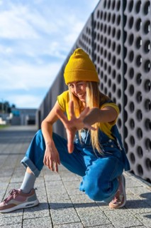 Winking blonde dancer with yellow woolen hat and blue dungarees performing urban dance outdoors