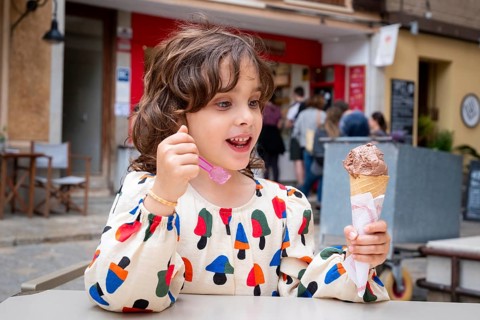 A cheerful child, with curly hair and a colorful dress, savors chocolate ice cream while sitting in
