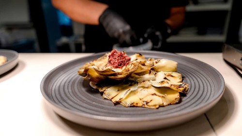A chef wears black gloves while preparing a deliciously plated artichoke dish topped with a vibrant