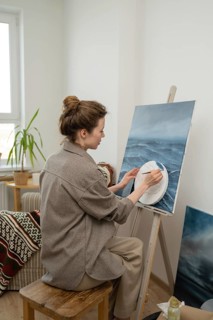 An artist sits on a wooden stool, painting a seascape on a canvas. Surrounded by art supplies, the