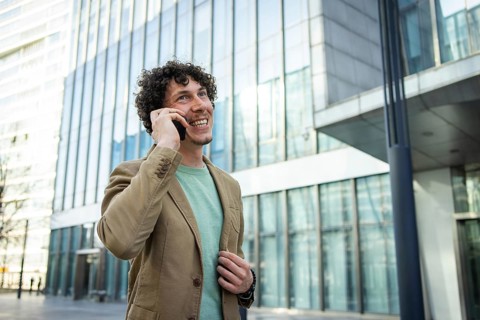 A cheerful man in a blazer is talking on his smartphone outside a sleek, glass paneled office