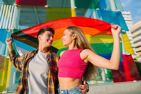 A joyful lesbian couple embraces while holding the Pride and Transgender flags. Their loving gaze