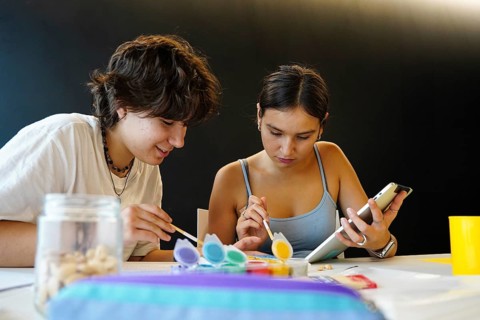 Two teenagers, a non-binary individual and a female, are deeply engaged in a creative art project