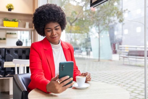 A stylish woman in a vibrant red blazer enjoys a cup of coffee at a cafe, looking at her smartphone