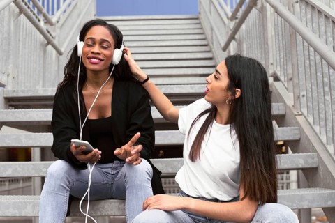 Two friends are sitting on outdoor stairs. One is listening to music with headphones, smiling while