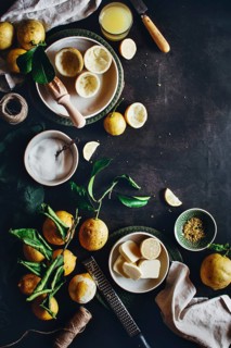 Fresh lemons with zest tool, a bowl of sugar, butter slices, and a glass of juice are arranged on a
