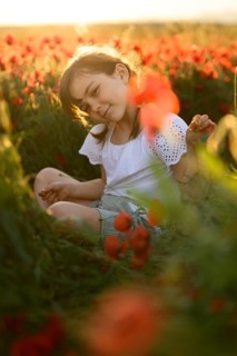A young girl smiles gently as she sits amidst a vivid field of red poppies (Papaver rhoeas),