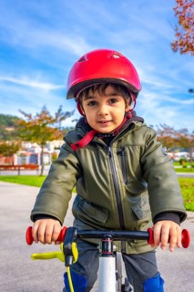 A young boy wearing a green jacket and a red helmet is riding a bike. The boy is smiling and he is