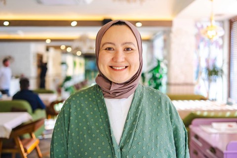 Asian woman wearing a green hijab smiling warmly in a cozy cafe. Her serene expression reflects