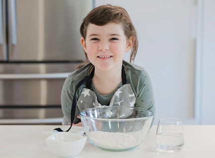 A cheerful child wearing an apron smiles while preparing to bake in a modern kitchen. The setup
