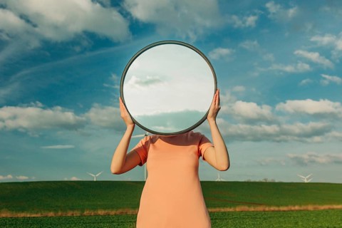 A person stands in a field, holding a circular mirror that reflects the blue sky and clouds,