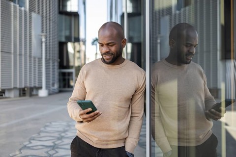 A cuban man in a beige sweater leans against a glass building facade, engrossed in his smartphone.