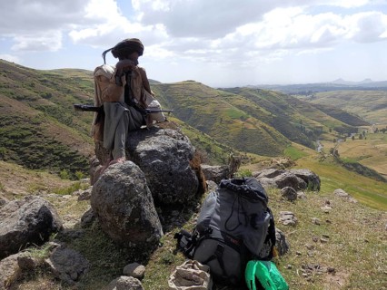 Man sitting with his rifle, Kalashnikov, on a rock with backpack in the foreground, surrounded by