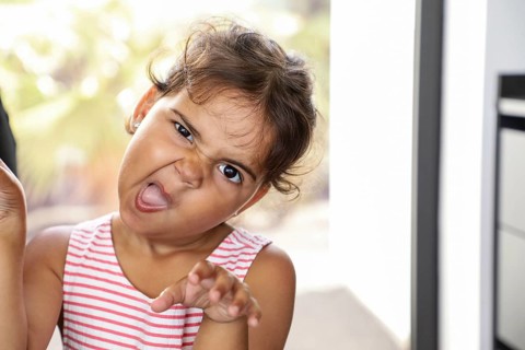 A playful child in a striped top strikes a silly face and gestures dramatically while indoors. The