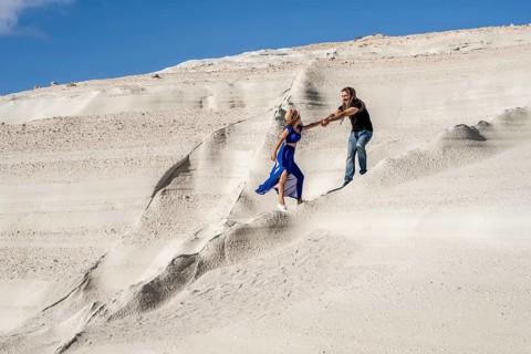 A man and woman joyfully hold hands and run down the unique white sandy slopes of Sarakiniko Beach