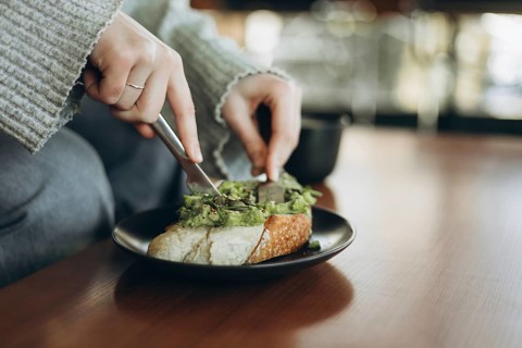 A person spreads fresh mashed avocado on a slice of crusty bread with a knife. The scene exudes a