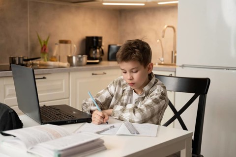 A young boy concentrates on his homework at a kitchen table. Surrounding him are a laptop and