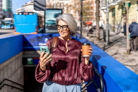 Woman standing on a busy city street near a subway entrance, observing content on her mobile phone