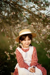 A young girl in a straw hat and red floral dress with a white apron sits under a blossoming almond