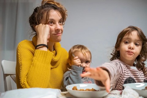 A mother, looking tired yet content, sits at a dining table with her two young children during
