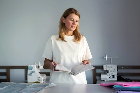 Woman in a bright sewing workshop, reviewing a pattern guide with sewing machines and colorful