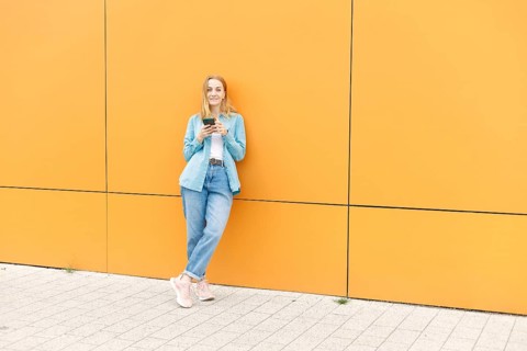 A woman dressed casually in denim holds a smartphone against a bright yellow wall in Warsaw She