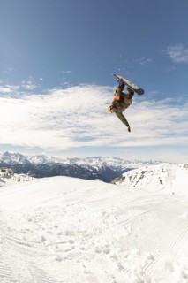 Dynamic photo of a snowboarder performing an aerial trick against a backdrop of snowy mountains.
