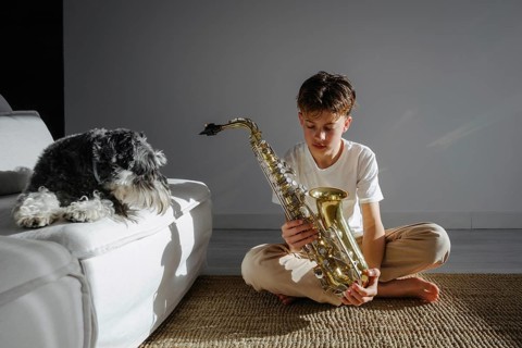 A young boy in a white shirt sits on a cushioned floor, intently studying a saxophone, with a