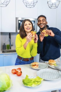Young multiethnic couple smiling and holding burgers they just made in their modern kitchen