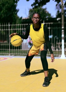 A woman athlete in a yellow vest practices dribbling on an outdoor basketball court. The scene
