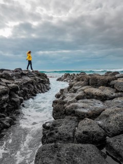 A woman in a yellow jacket stands on rocky terrain by the ocean at Timanfaya National Park,