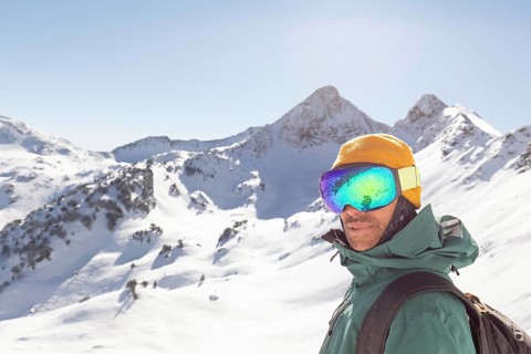 A snowboarder dressed in winter gear stands confidently in a backcountry mountain setting. The