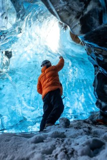 A man in an orange jacket is standing in a blue cave. The cave is filled with ice and the man is