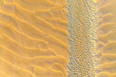 An aerial photograph showcases vast, golden sand dunes and intricate textures in a desert landscape
