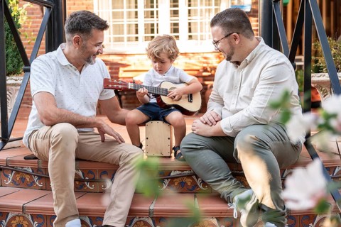 A joyful gay couple shares a musical moment with their young son, who is playing a small guitar.