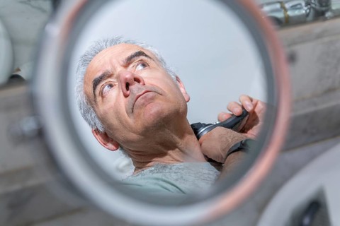 An older man is seen from the mirror's perspective, carefully using an electric razor. The image