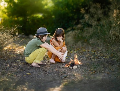 Two children sit by a small campfire in a woodland area. They are enjoying the outdoors, surrounded